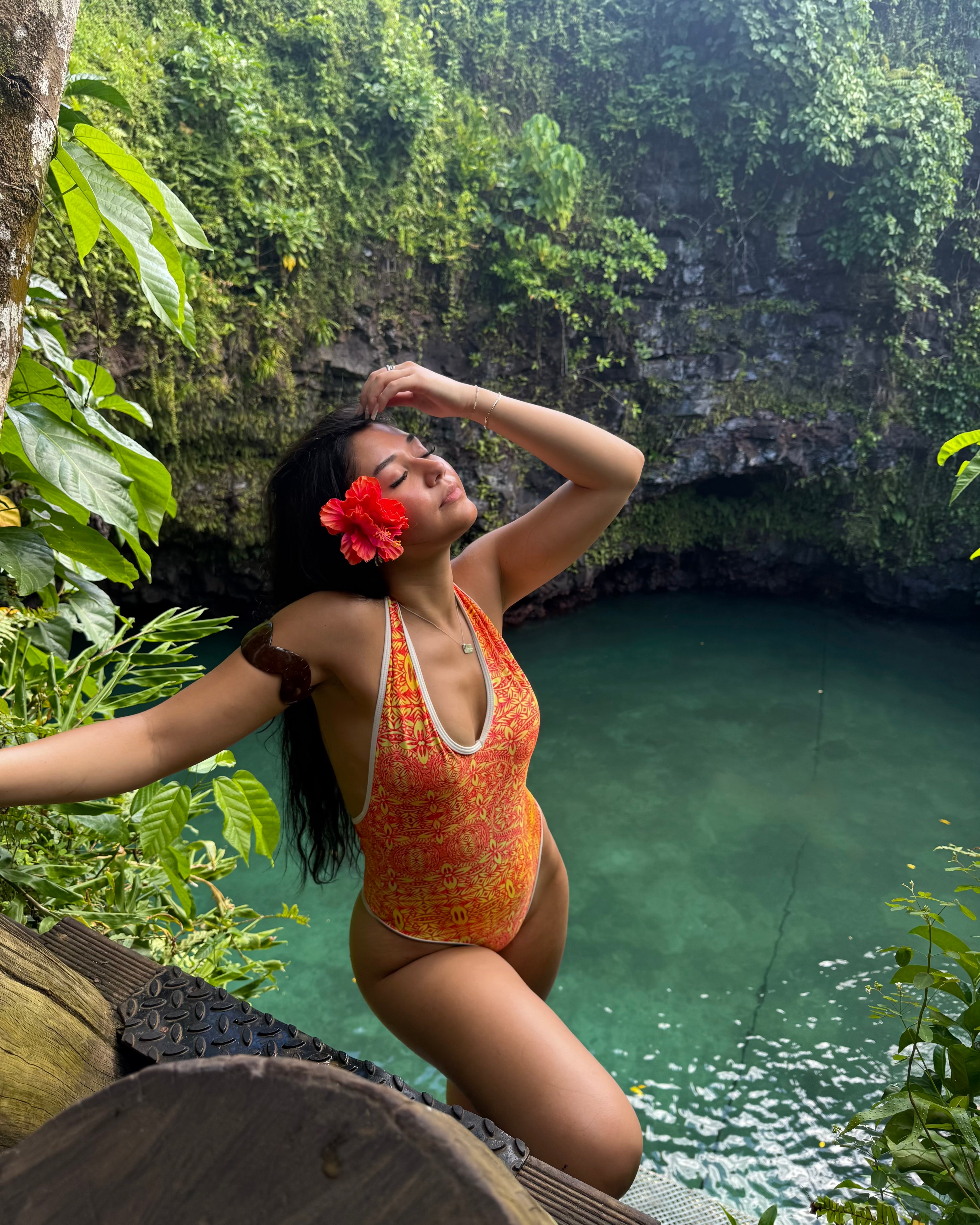 Woman in an orange swimsuit with a red flower in her hair, standing by a natural pool surrounded by greenery.